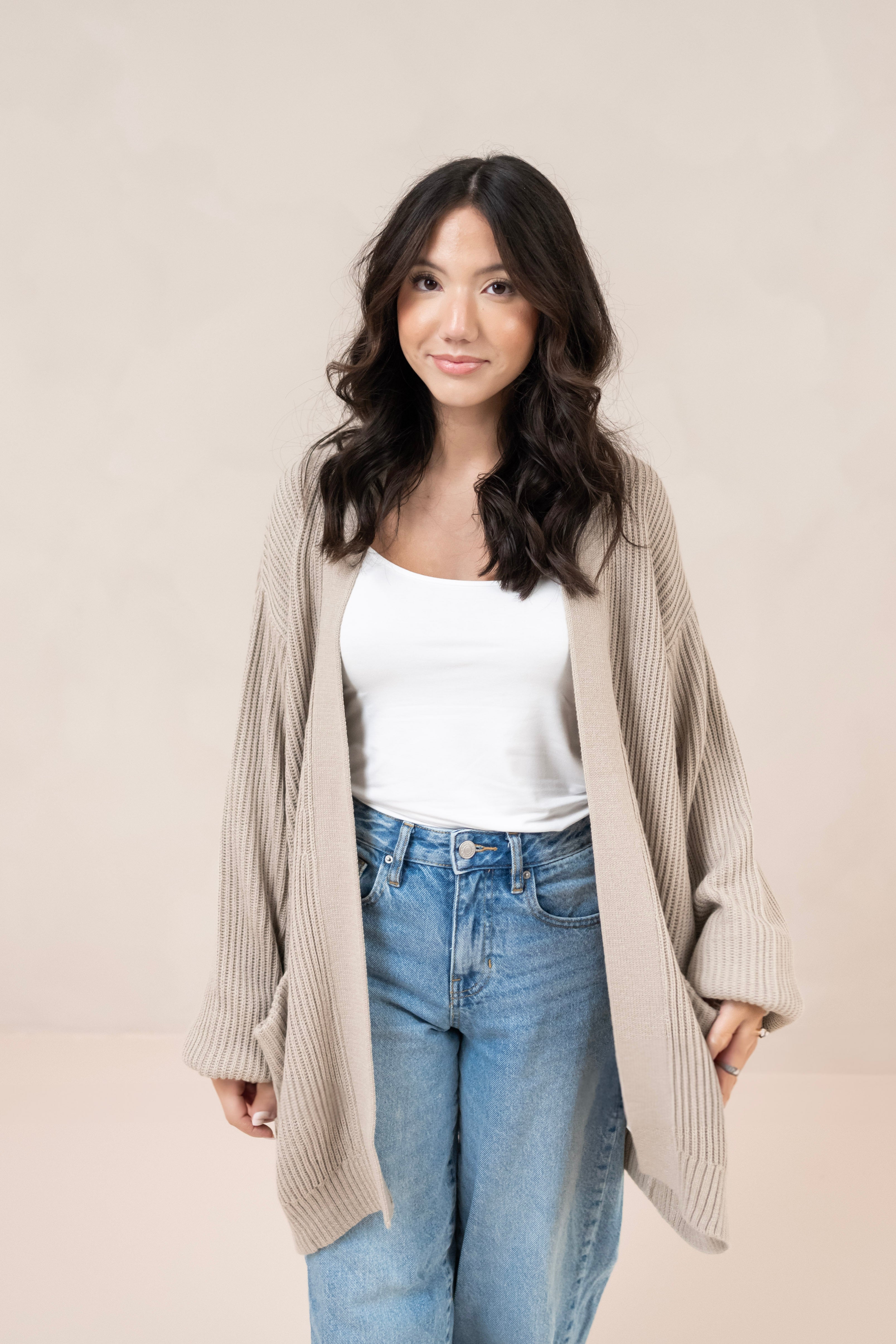 Woman wearing a tan cardigan, white top, and blue jeans against a plain background