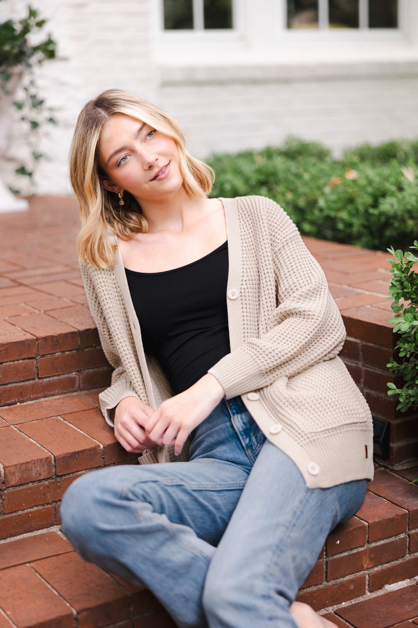 Modal sitting outdoors wearing Chunky Knit Women's Waffle Cardigan in Almond over midnight camisole and blue jeans
