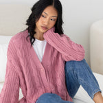 Woman wearing a pink knitted cardigan and blue jeans sitting on a bed.   ::: Model is 5'6" wearing size XS ///