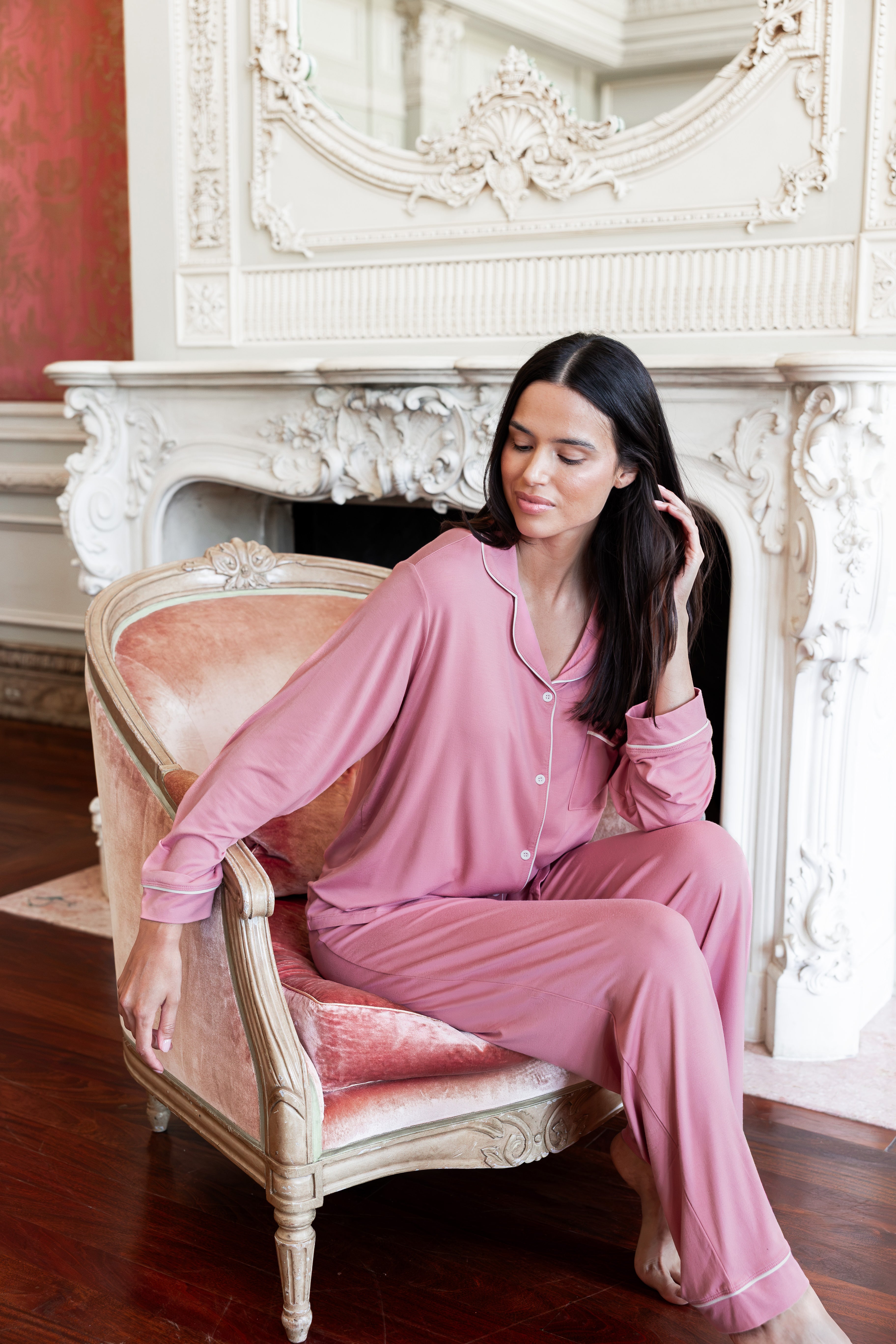 A woman wearing a dusty pink pajama set sits on an ornate pink velvet armchair in front of a white fireplace mantel.
---1605 1