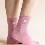 A close-up of a person's feet wearing light pink ribbed crew socks with a small white geometric logo on the side, set against a solid pale beige background.