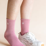 A close-up of a person's feet wearing pink crew socks and a white athletic sneaker against a solid light beige background.