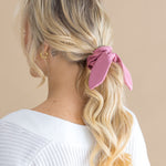 Woman with blonde hair styled in a ponytail with a pink scrunchie, wearing a white top against a beige background.