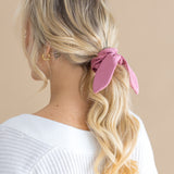 Woman with blonde hair styled in a ponytail with a pink scrunchie, wearing a white top against a beige background.