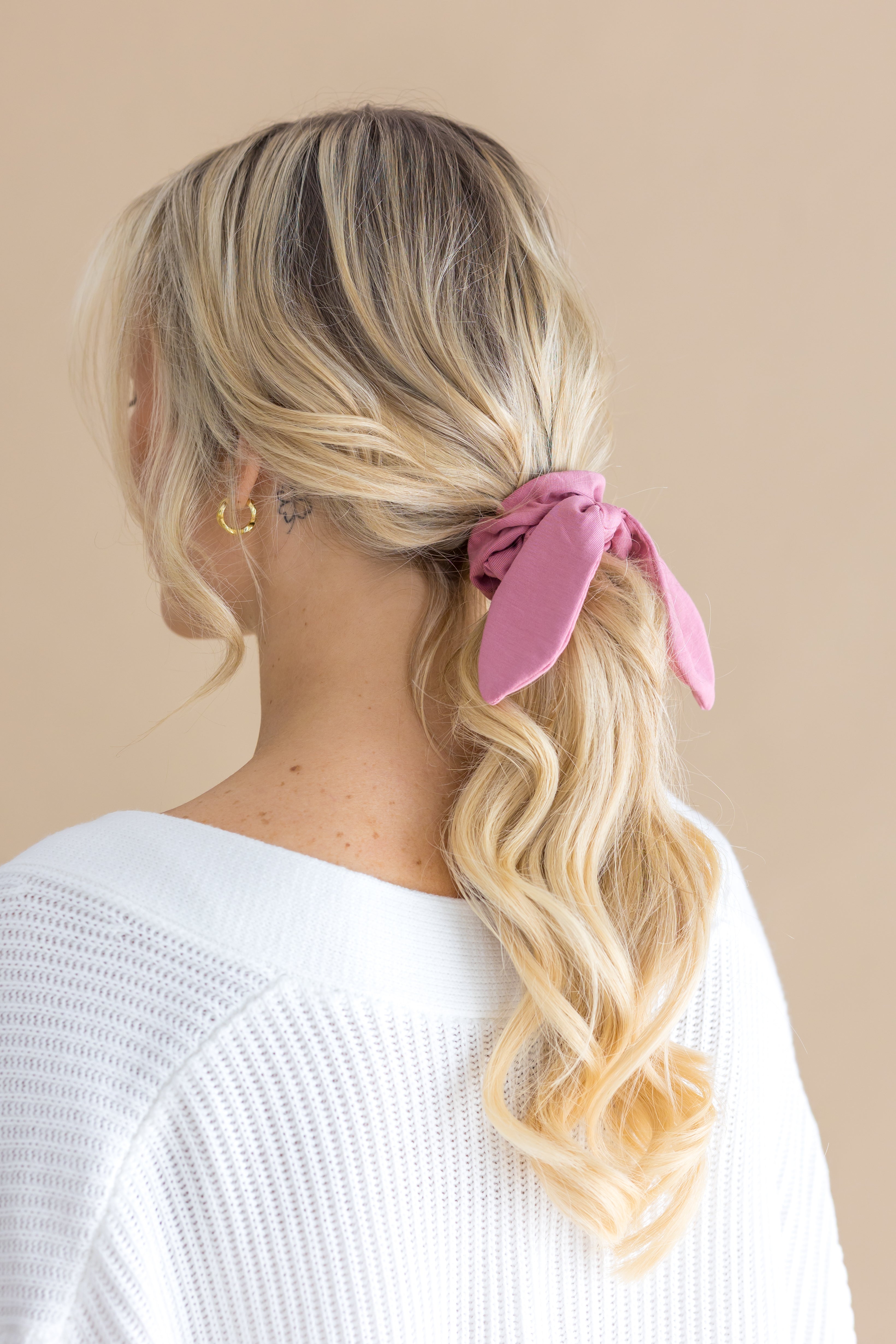 Woman with blonde hair styled in a ponytail with a pink scrunchie, wearing a white top against a beige background.