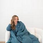 Smiling model sitting on a white chair while wrapped in the Quilt Blanket in Atlantic 2.5 TOG