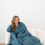 Smiling model sitting on a white chair while wrapped in the Quilt Blanket in Atlantic 2.5 TOG