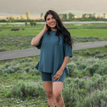 Smiling model wearing Kyte Biker Short Set in Atlantic paired with cowboy boots, standing in a grassy field with mountains in the background
