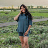 Smiling model wearing Kyte Biker Short Set in Atlantic paired with cowboy boots, standing in a grassy field with mountains in the background
