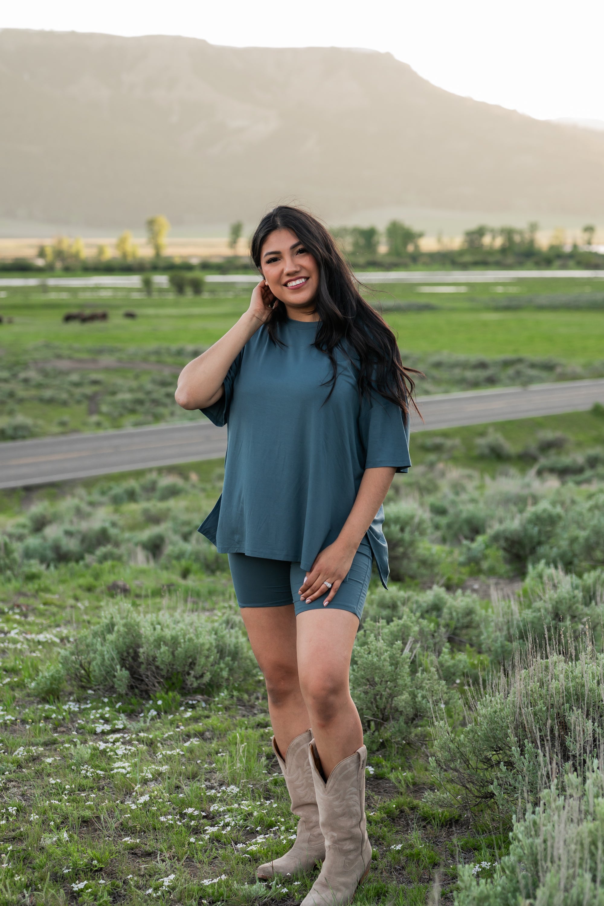Smiling model wearing Kyte Biker Short Set in Atlantic paired with cowboy boots, standing in a grassy field with mountains in the background