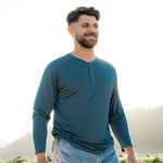 Model wearing Men's Long Sleeve Henley in Atlantic and light blue jeans, standing outdoors in a grassy area with the sun shining behind him, smiling and looking off into the distance.