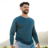 Model wearing Men's Long Sleeve Henley in Atlantic and light blue jeans, standing outdoors in a grassy area with the sun shining behind him, smiling and looking off into the distance.