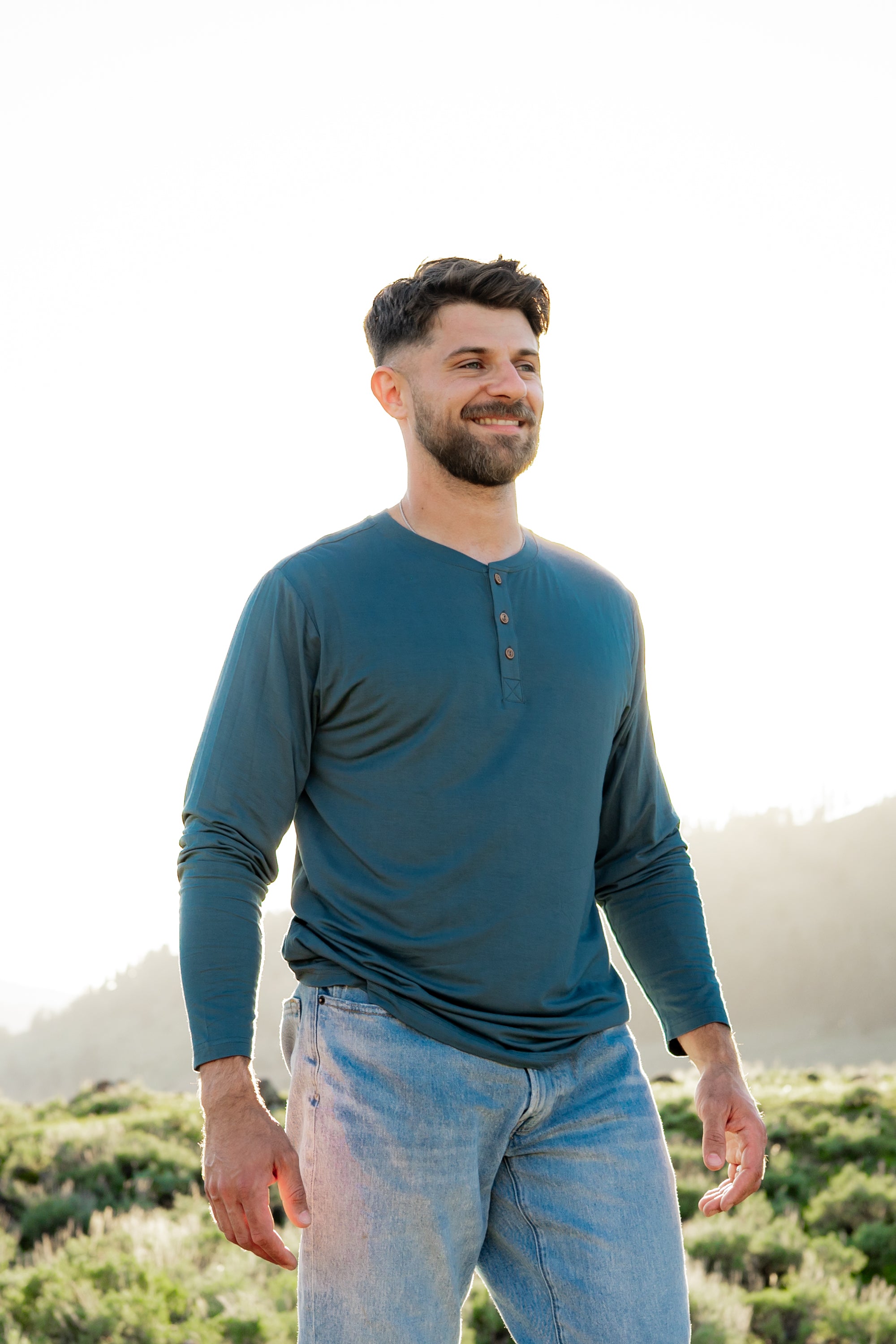 Model wearing Men's Long Sleeve Henley in Atlantic and light blue jeans, standing outdoors in a grassy area with the sun shining behind him, smiling and looking off into the distance.