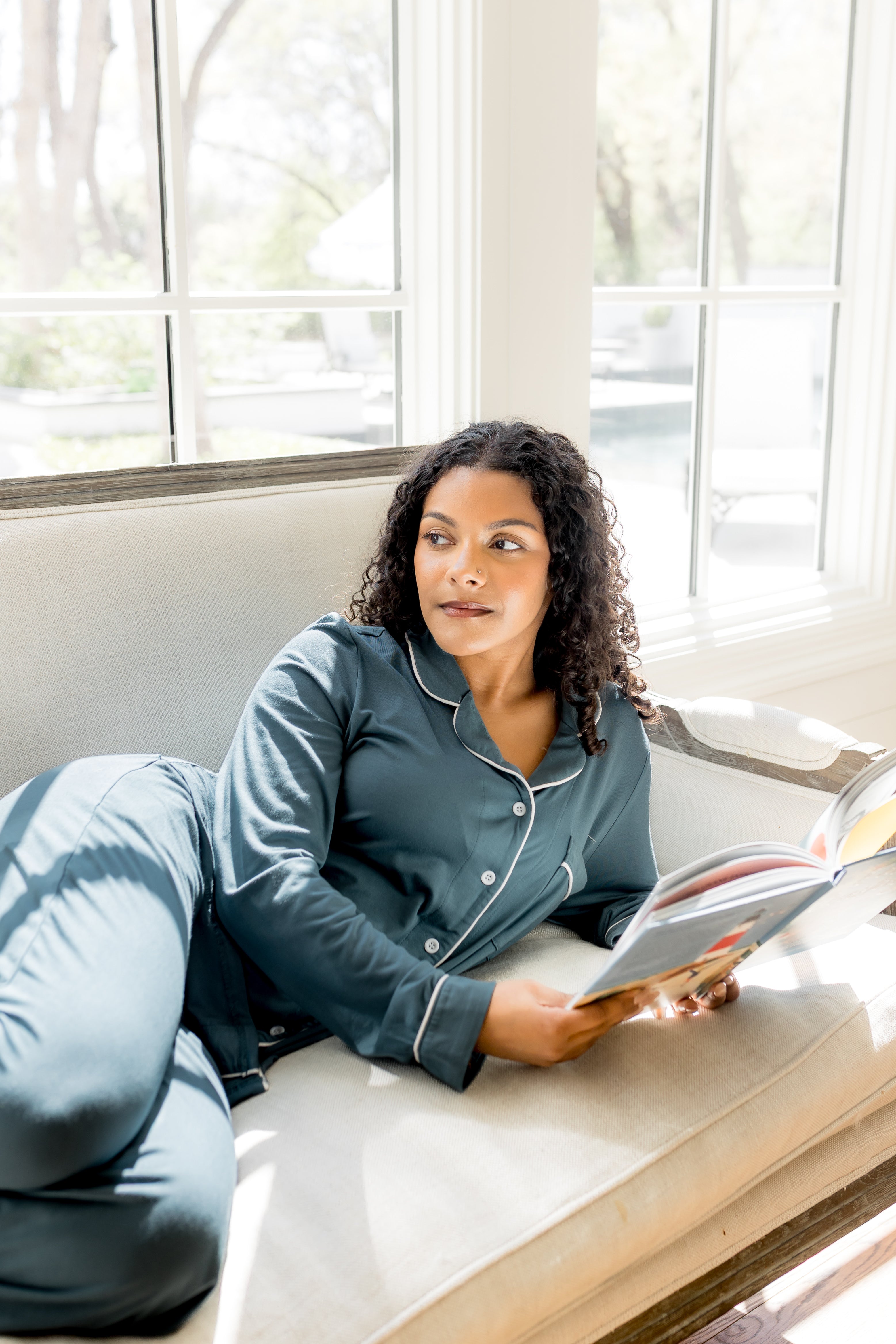 Woman in blue pajamas reading a book on a couch by a window