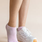 A close-up of a person's legs wearing a pair of light pink low-cut athletic socks and white sneakers with orange soles, set against a solid beige background.