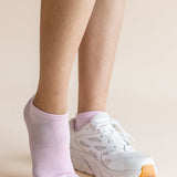A close-up of a person's legs wearing a pair of light pink low-cut athletic socks and white sneakers with orange soles, set against a solid beige background.