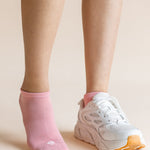 A close-up of a person's lower legs and feet, showing one ankle in a light pink no-show sock and the other foot wearing a white athletic sneaker with a pink collar and orange outsole, against a solid pale beige background.