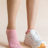 A close-up of a person's lower legs and feet, showing one ankle in a light pink no-show sock and the other foot wearing a white athletic sneaker with a pink collar and orange outsole, against a solid pale beige background.
