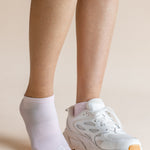 A person's legs and feet shown wearing a pair of white ankle socks, with one foot in a white sneaker and the other barefoot, against a solid beige background.
