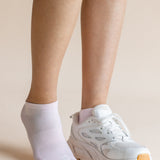 A person's legs and feet shown wearing a pair of white ankle socks, with one foot in a white sneaker and the other barefoot, against a solid beige background.