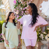 Two women standing outdoors with floral decorations in the background wearing the Biker Short Sets in Thistle and Basil
