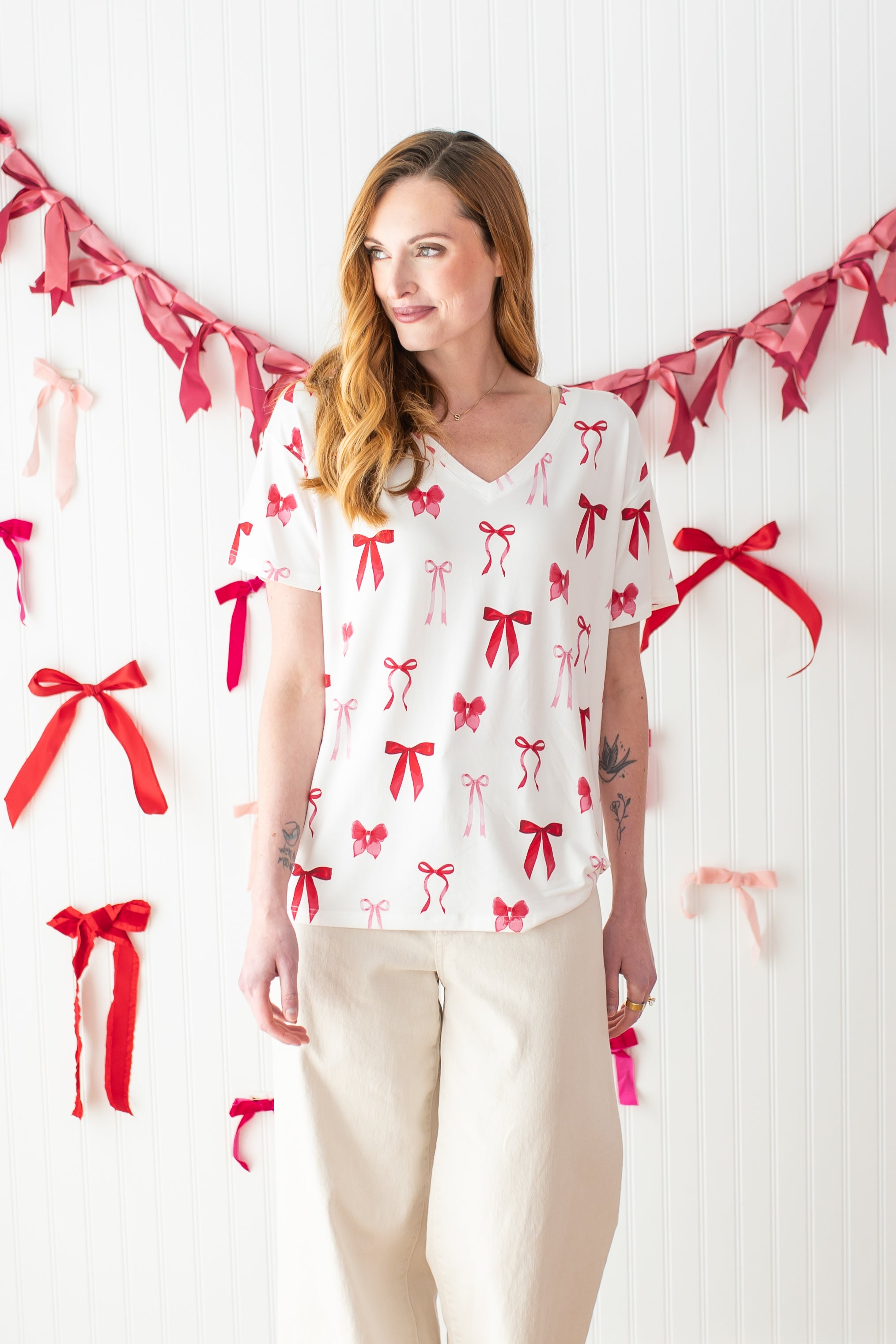 Model standing in front of a white wall decorated with bows and wearing the Bamboo Women's Relaxed Fit V-Neck in Big Love Bow