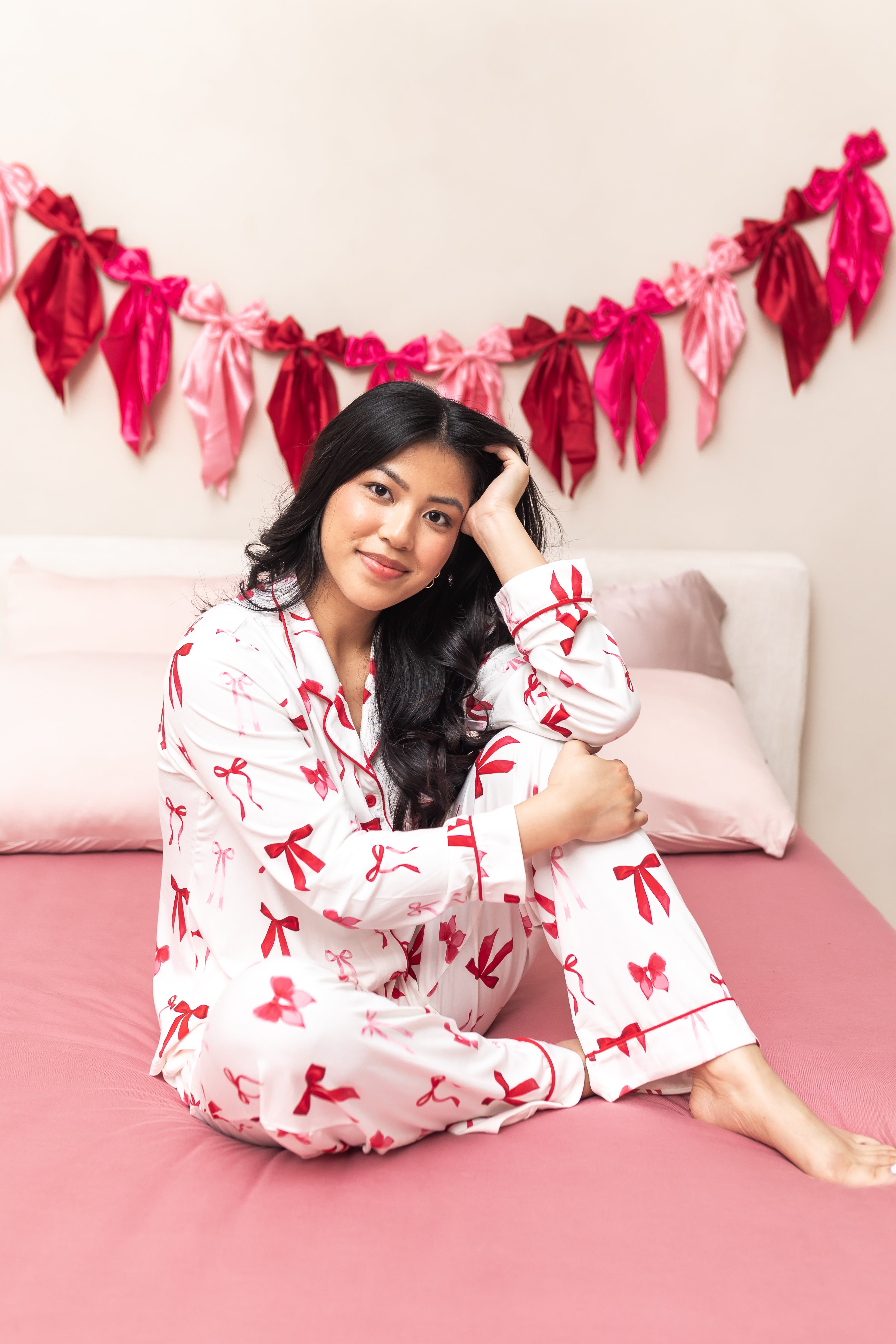 Woman sitting on a pink bed while wearing the Bamboo Long-Sleeved Women's Pajama Set in Big Love Bow