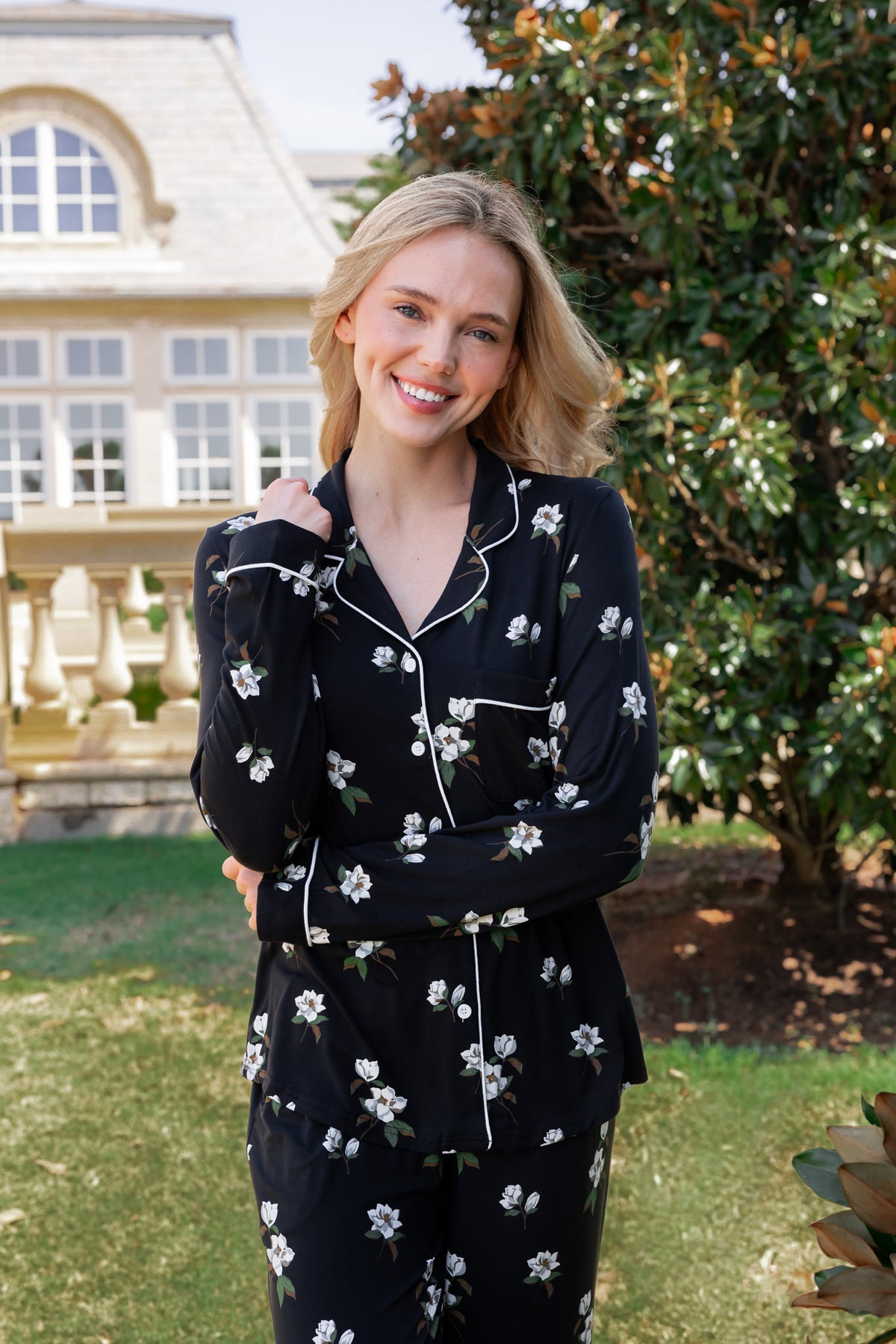 A woman stands outdoors in a garden wearing a black long-sleeve pajama set with a white floral print and white piping. The background shows green grass, a large tree, and a house.