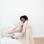 Smiling model sitting on a white couch while wrapped in a Blanket in Bisque 