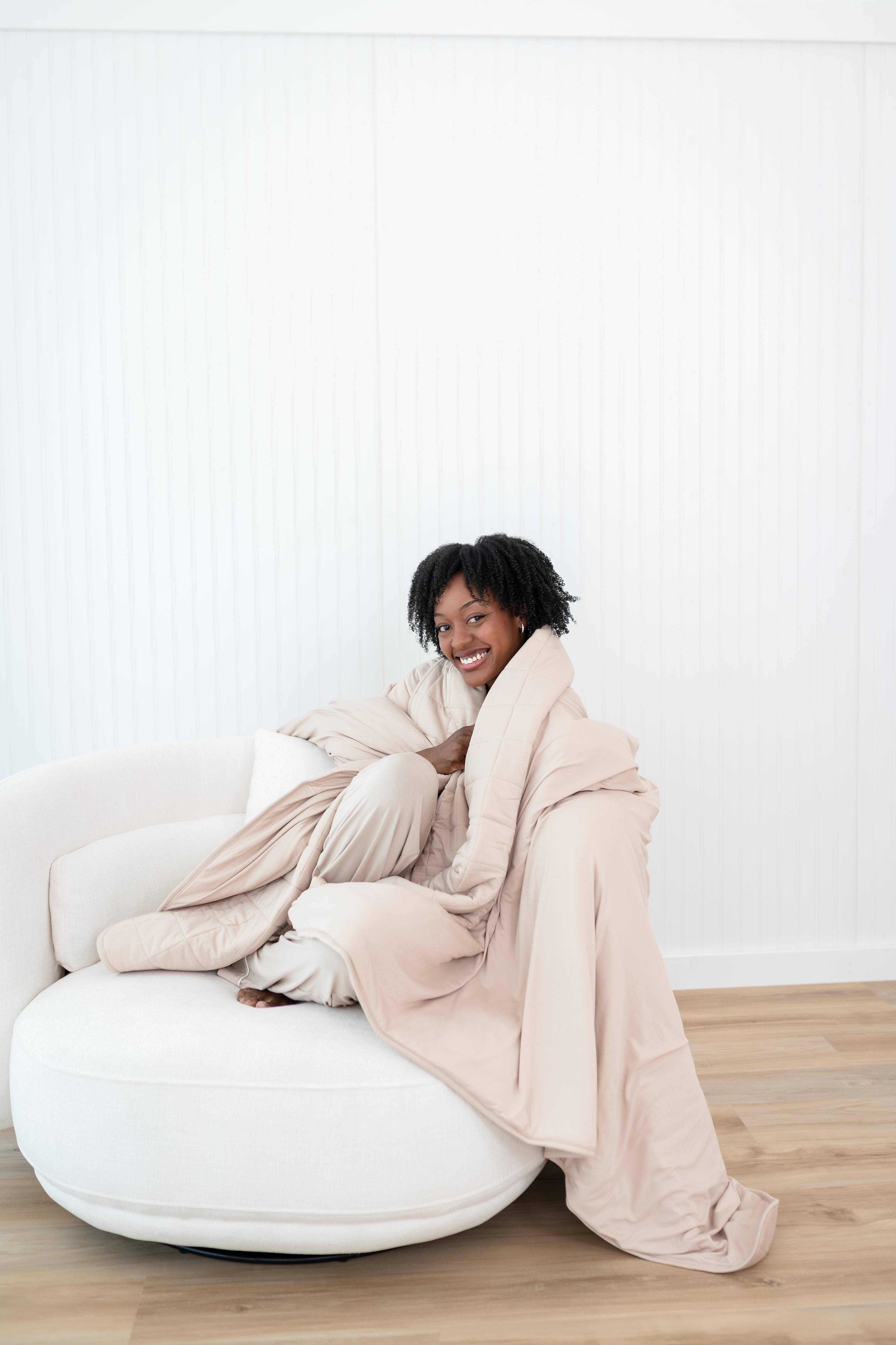 Smiling model sitting on a white couch while wrapped in a Blanket in Bisque 