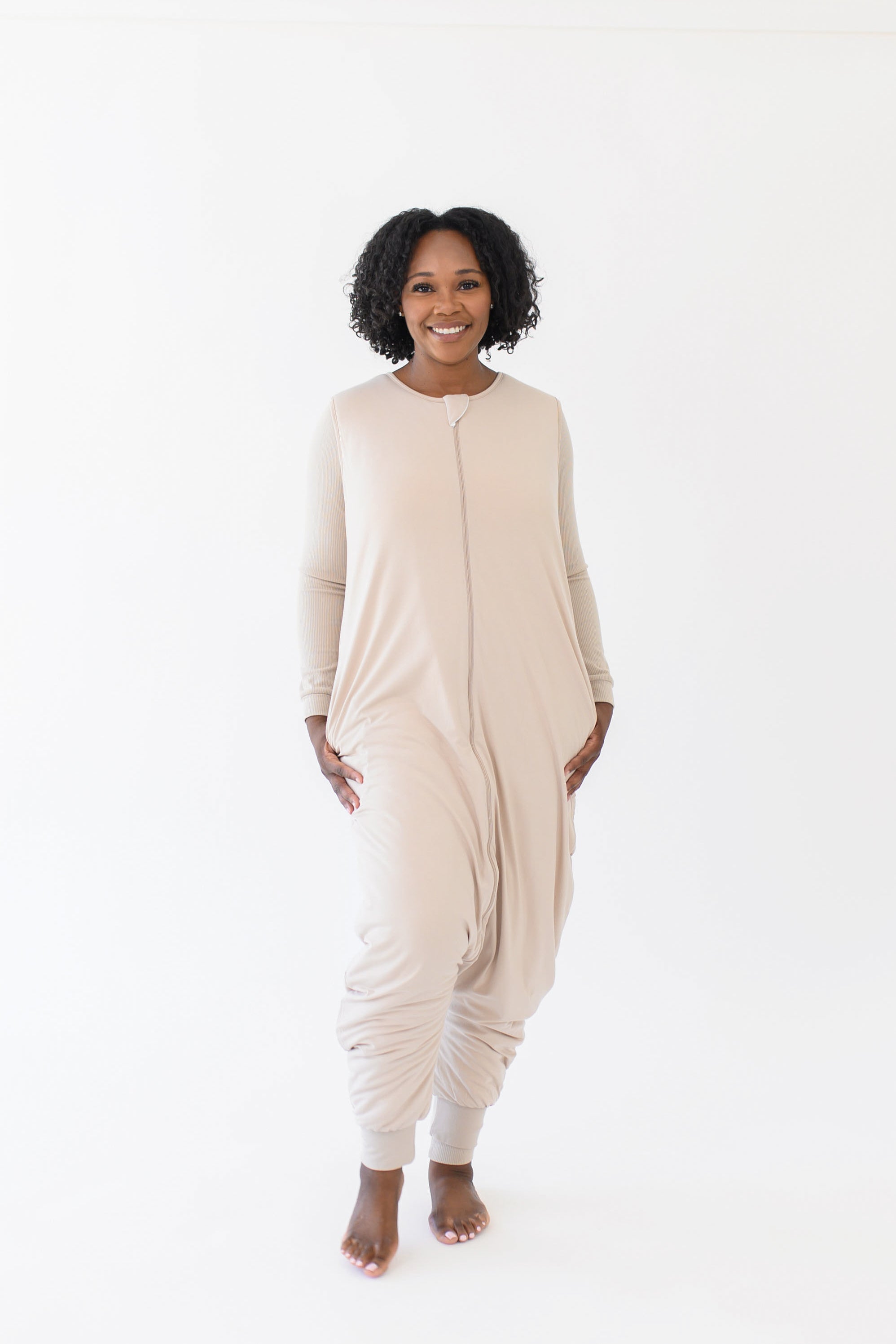 Smiling model standing in a white room while wearing the Kyte Adult Sleep Bag Walker in Bisque