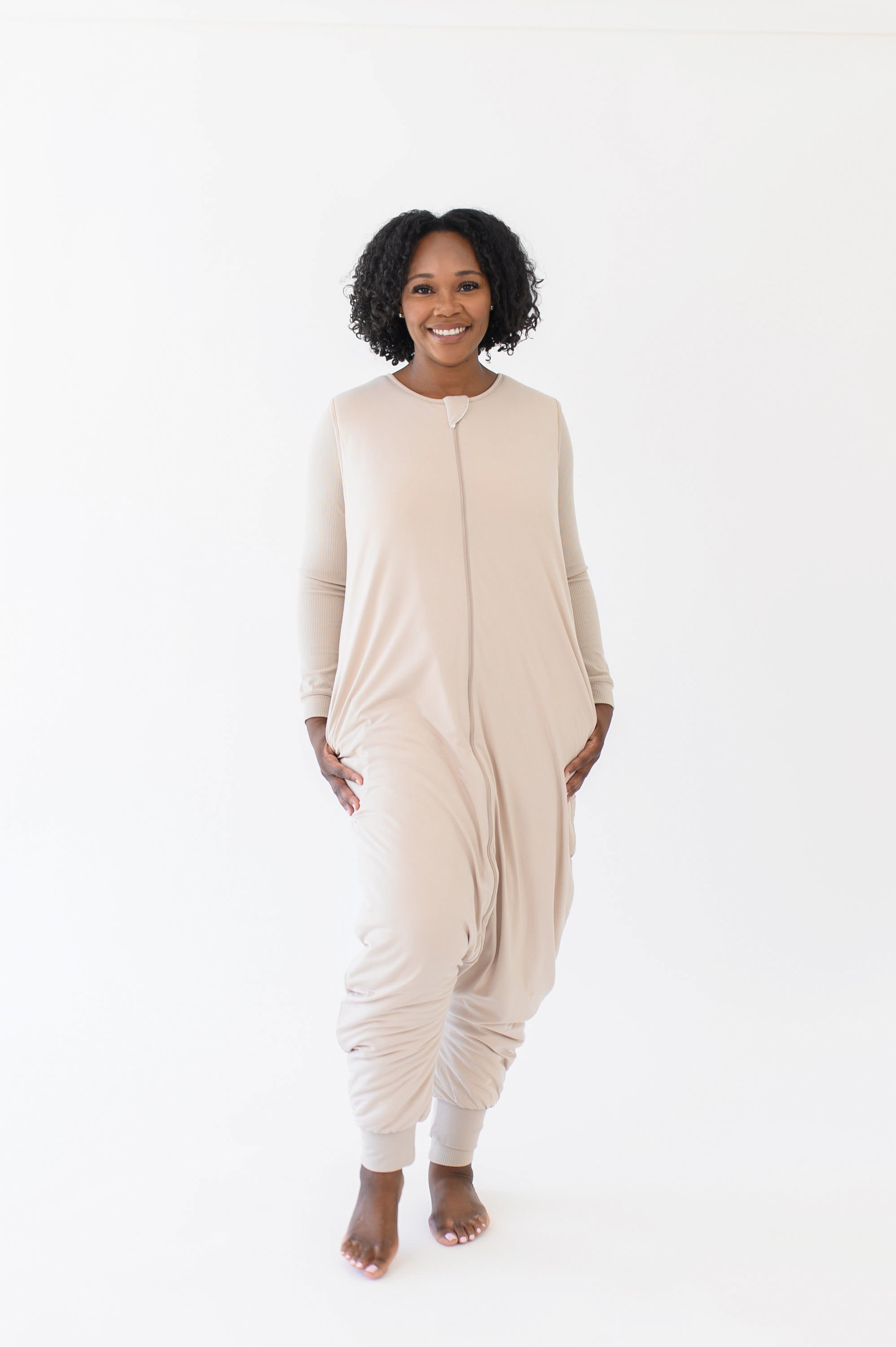 Smiling model standing in a white room while wearing the Kyte Adult Sleep Bag Walker in Bisque