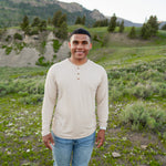 Smiling male model standing in grassy field while waring Men's Long Sleeve Henley in Bisque and light washed jeans
