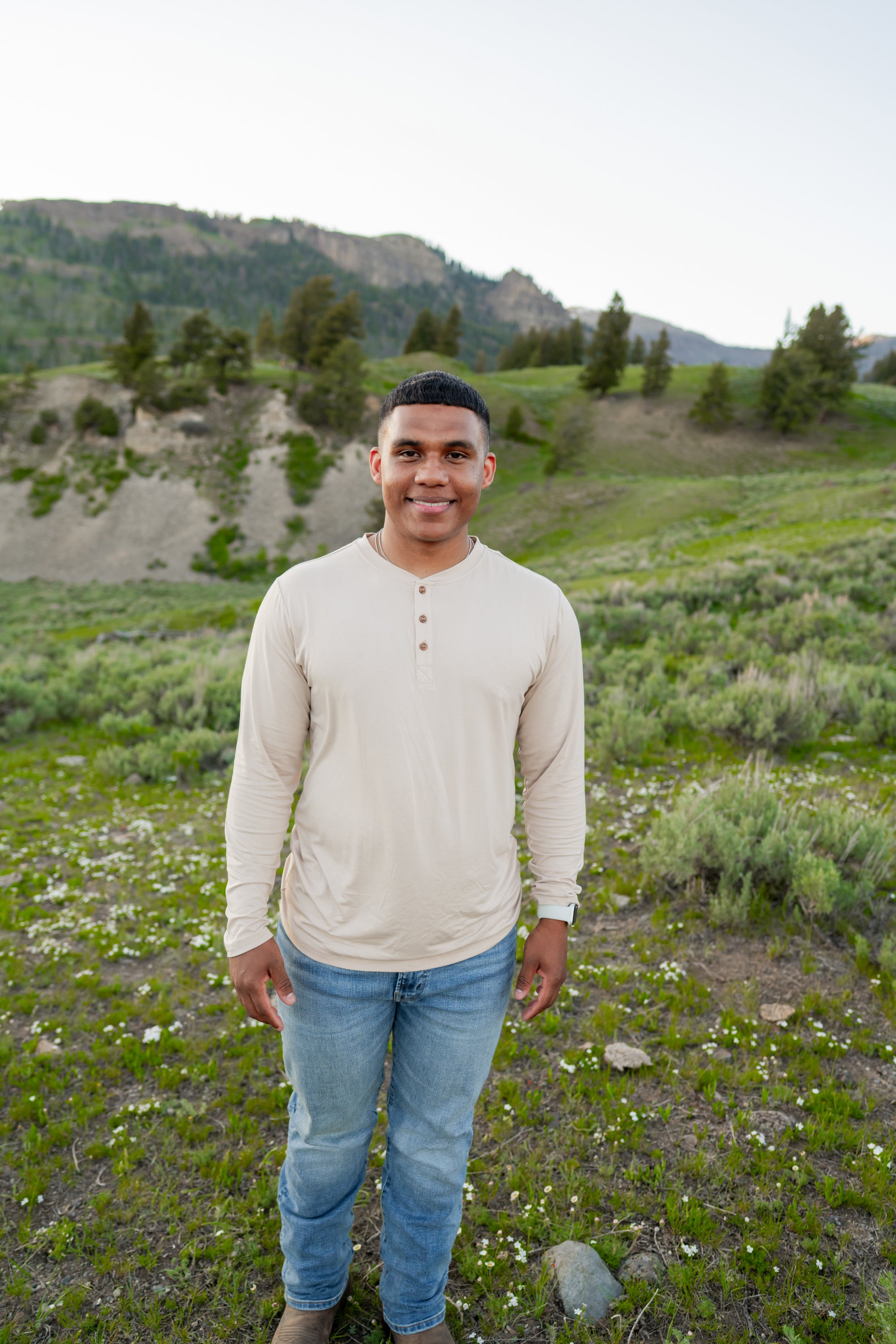Smiling male model standing in grassy field while waring Men's Long Sleeve Henley in Bisque and light washed jeans