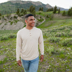 Model walking in grassy field with mountains in background while wearing Men's Long Sleeve Henley in Bisque