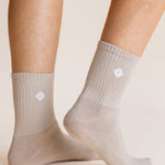A close-up of a person's feet and lower legs wearing light beige crew socks with a small white geometric logo on the side, set against a solid warm beige background.