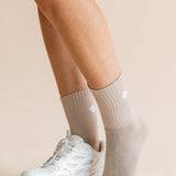 A close-up of a person's legs wearing light beige ribbed crew socks and a white athletic sneaker against a solid beige background.