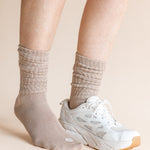 A close-up of a person's legs wearing beige ribbed crew socks and a white athletic sneaker against a solid light beige background.