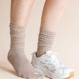 A close-up of a person's legs wearing beige ribbed crew socks and a white athletic sneaker against a solid light beige background.