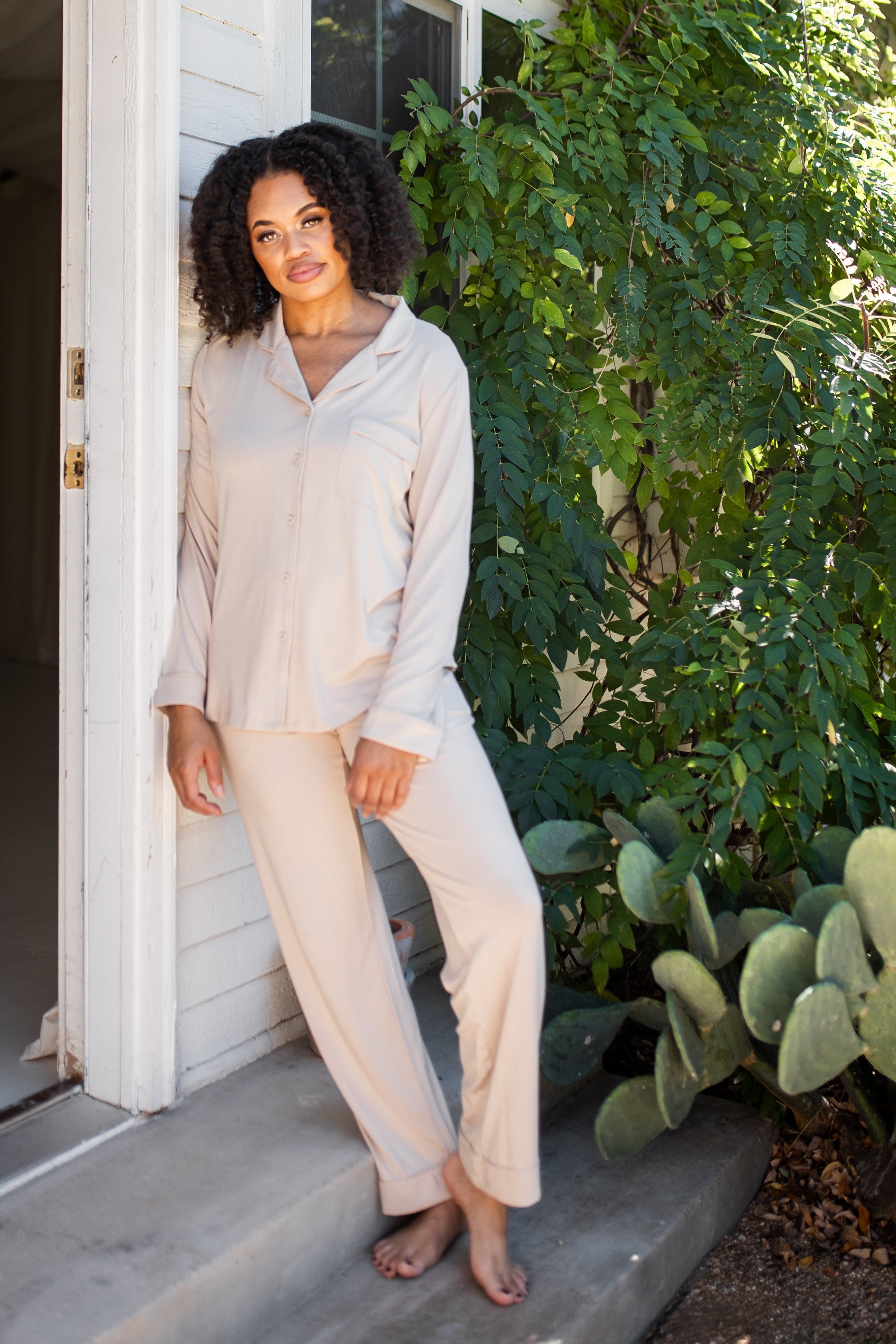 Female model leaning against a white house wearing the Long-Sleeved Women's Pajama Set in Bisque with Bisque Trim