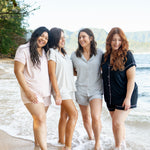 Multiple models wearing different colors of Kyte Baby's Women's Short Sleeve Pajama Sets on the beach with their feet in the ocean