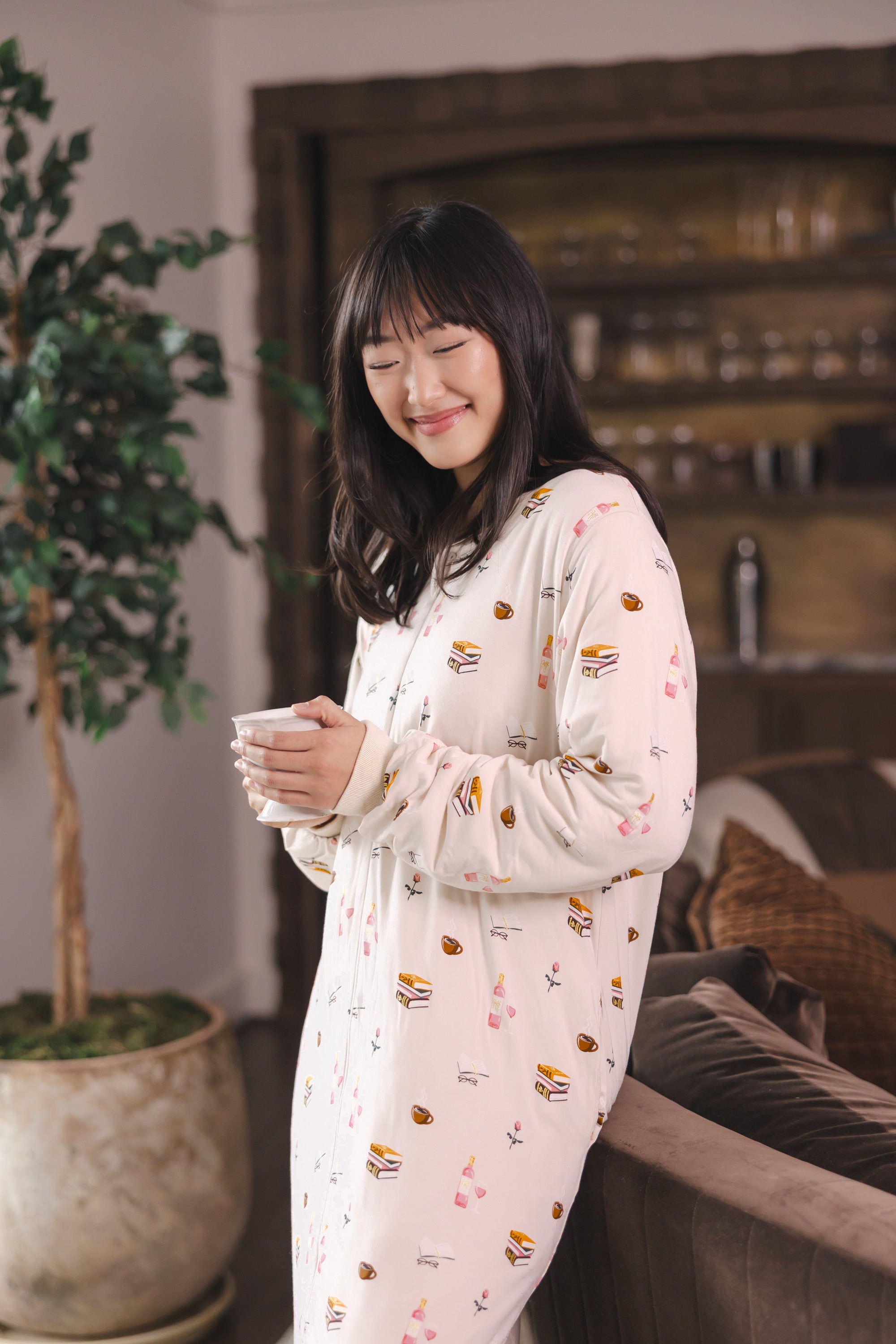 Modal smiling in a living room holding a cup wearing Beige Long Sleeve Adult Sleep Bag Walker in Book Club print