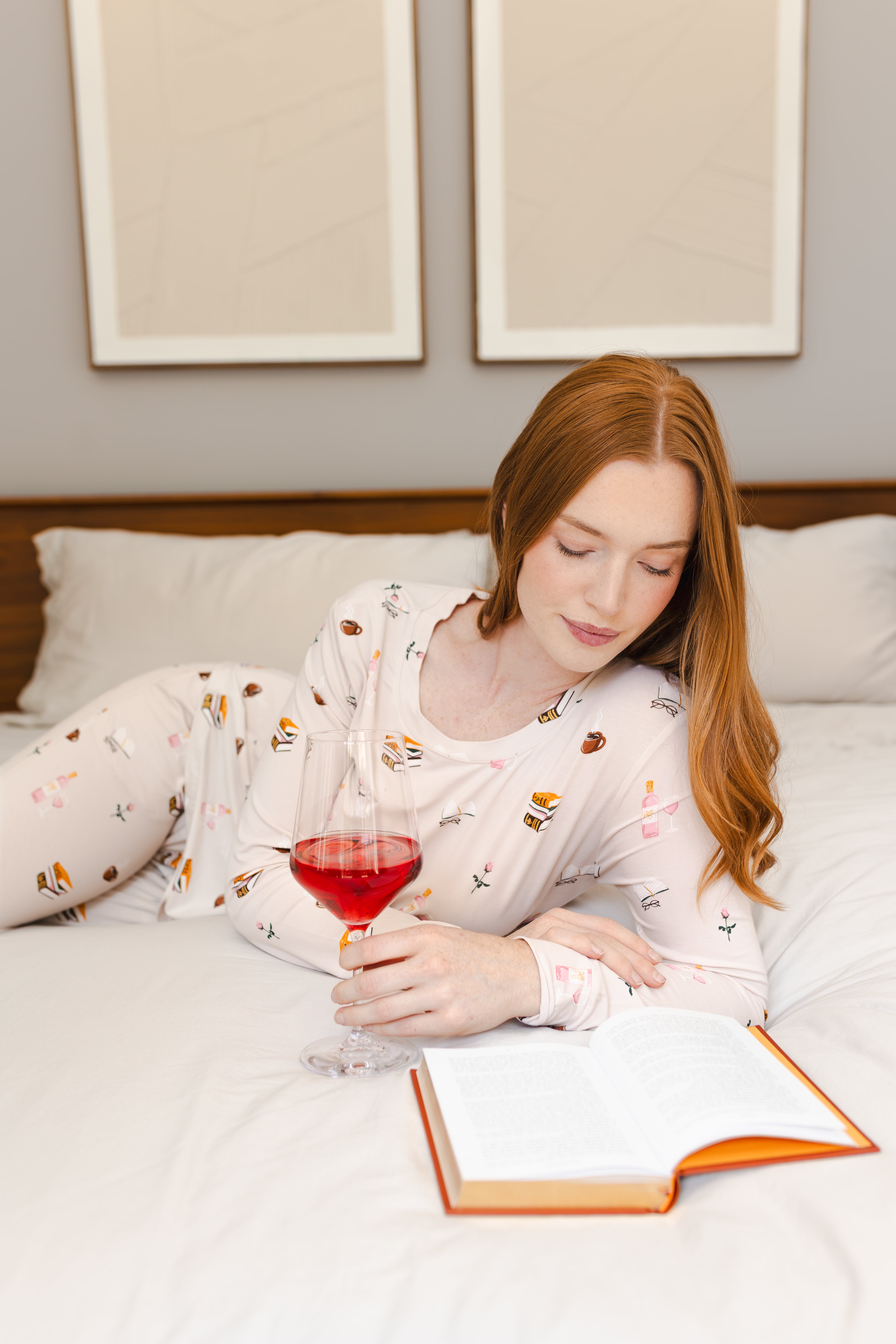 Modal wearing Women's Jogger Pajama Set in Book Club print lying on a bed reading a book with a drink in her hand