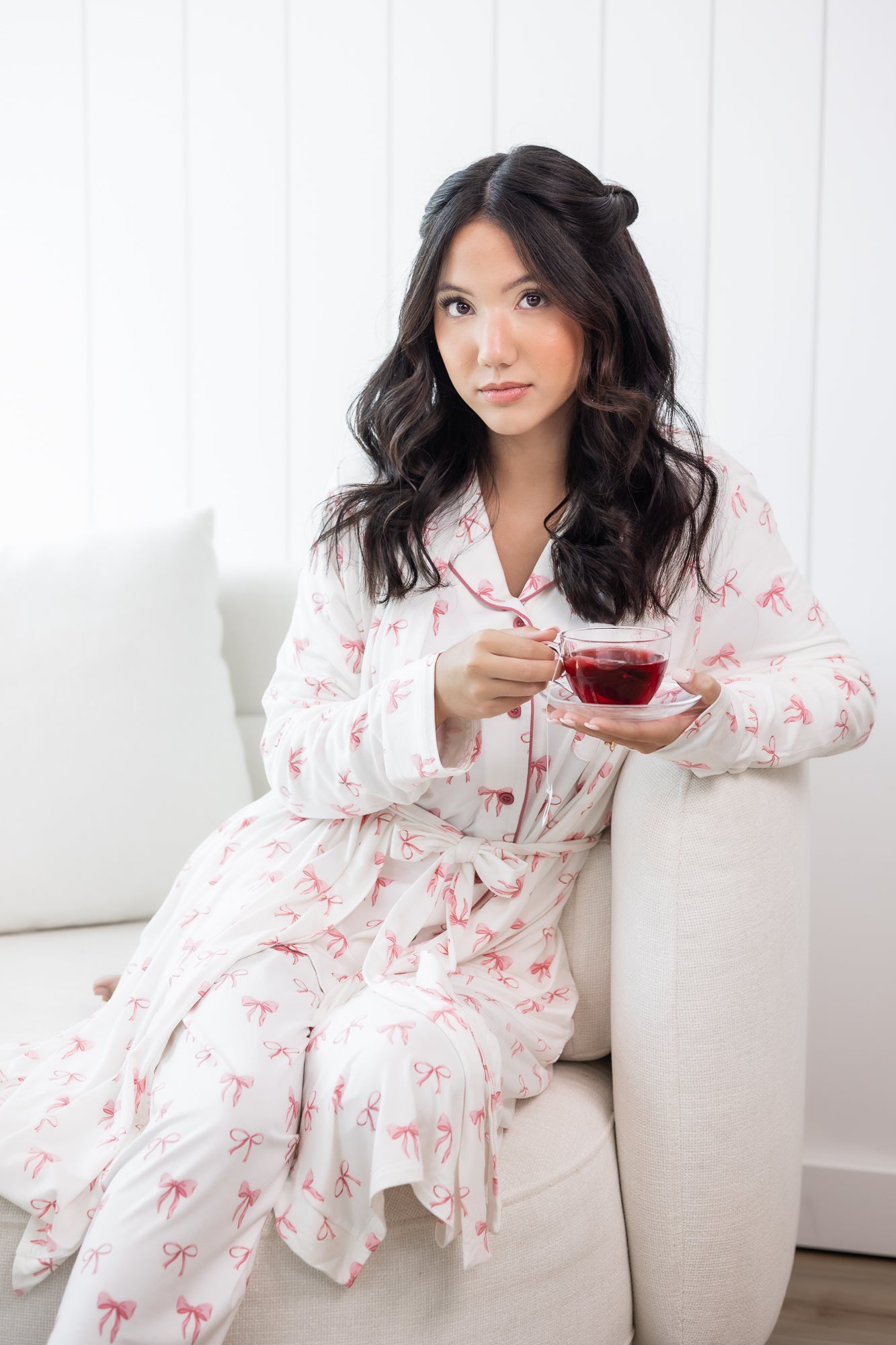 Woman in a white robe with pink bow pattern sitting on a white couch holding a glass of tea