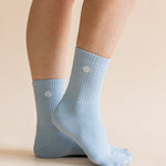 A pair of light blue crew socks worn on a person's feet, shown against a plain beige studio background. A small white geometric logo is visible on each sock.