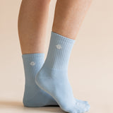 A pair of light blue crew socks worn on a person's feet, shown against a plain beige studio background. A small white geometric logo is visible on each sock.