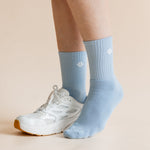 A person's legs and feet wearing light blue crew socks and a white athletic sneaker, shown against a solid pale beige background.