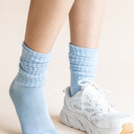 A close-up of a person's legs wearing light blue ribbed crew socks and a white athletic sneaker against a plain beige background.