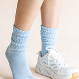 A close-up of a person's legs wearing light blue ribbed crew socks and a white athletic sneaker against a plain beige background.