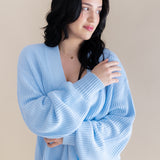 Female model wrapped in the Chunky Knit Women's Oversized Cardigan in Breeze standing in front of a light beige background
